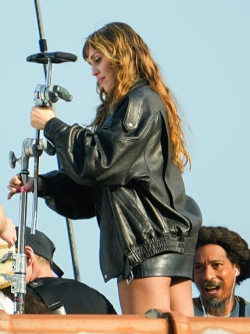 Person wearing a black leather jacket on a film set with equipment and other people in the background.