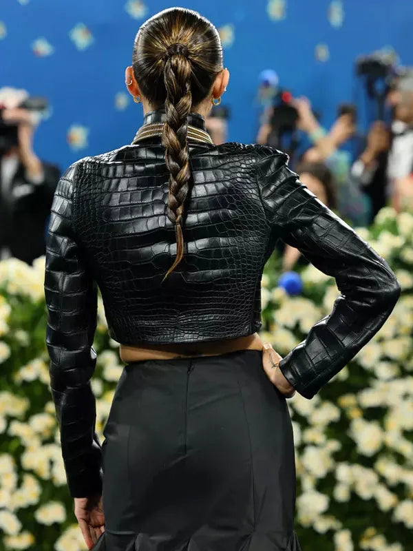 Person wearing a black leather jacket with a braided hairstyle, standing in front of a floral arrangement and blurred people.