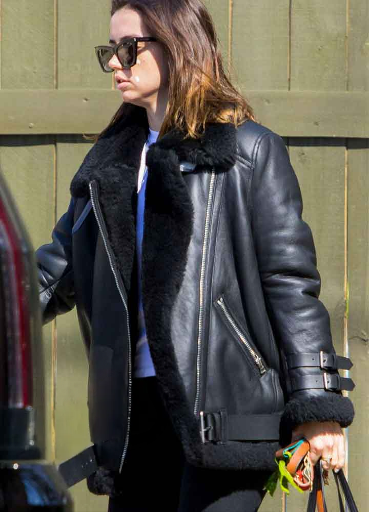 Person wearing a black leather jacket with fur trim, standing against a green wooden wall.